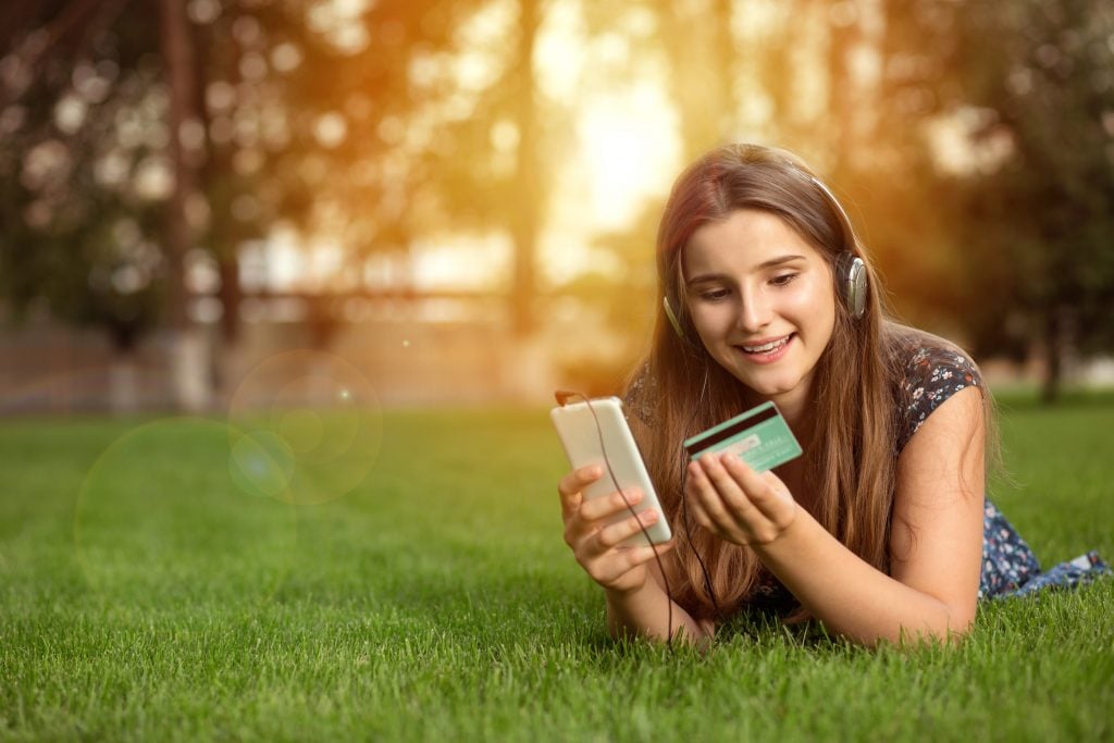 Portrait of a happy woman buying online with a smart phone outdoors lying down on green lawn headphones on head. Multicultural model, mixed race, asian russian girl. Copy space. People and technology.