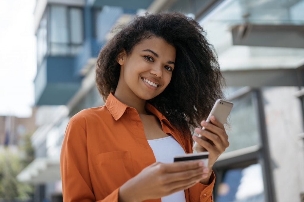 African American woman holding credit card, using smartphone with mobile application for online shopping with low prices and cash back. Happy hipster girl ordering food online, making transaction 