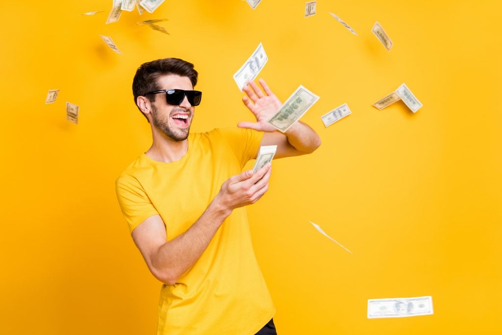 Photo of young handsome careless guy throwing money banknotes away, wealthy person wear sun specs casual t-shirt isolated bright yellow color background