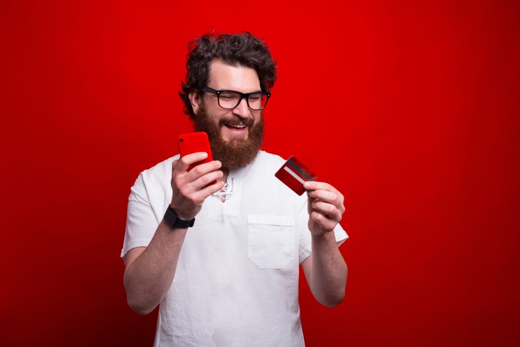 Happy smiling bearded hipsteris using phone and his credit card over red background