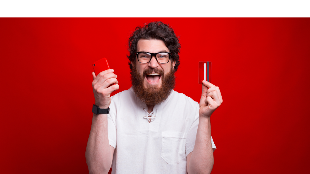 Excited bearded man is holding a phone and credit card over red background.