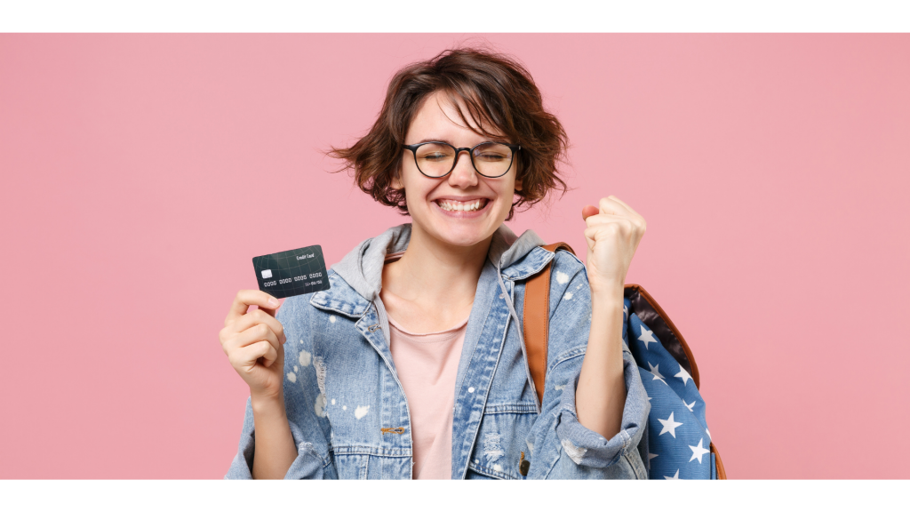 Happy young woman student in denim clothes glasses backpack posing isolated on pastel pink background. Education in high school university college concept. Doing winner gesture, hold credit bank card