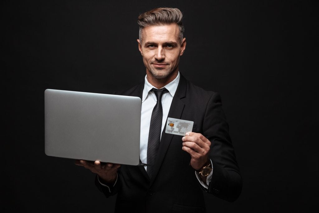Confident businessman wearing suit standing isolated over black background, holding laptop computer, showing plastic credit card