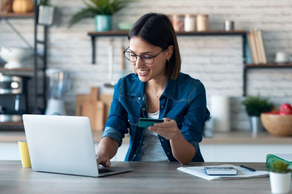 Young woman holding white credit card for shopping online with computer while sitting in the kitchen at home (apply Petal® 1 “Cash Back, No Fees” Visa® card).