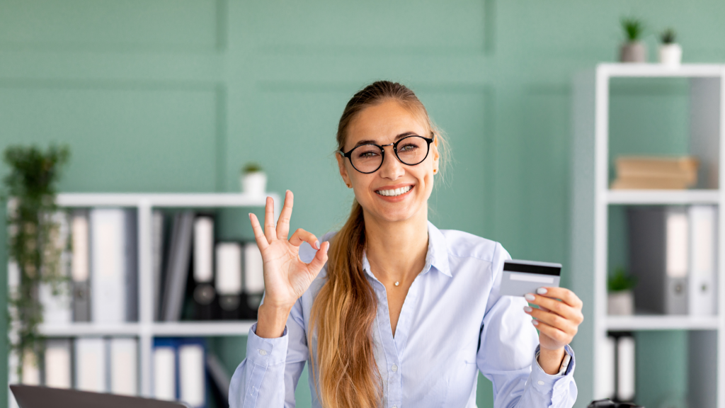 Accountant banker lady holding credit card and showing ok sign while sitting at workplace, smiling at camera in office