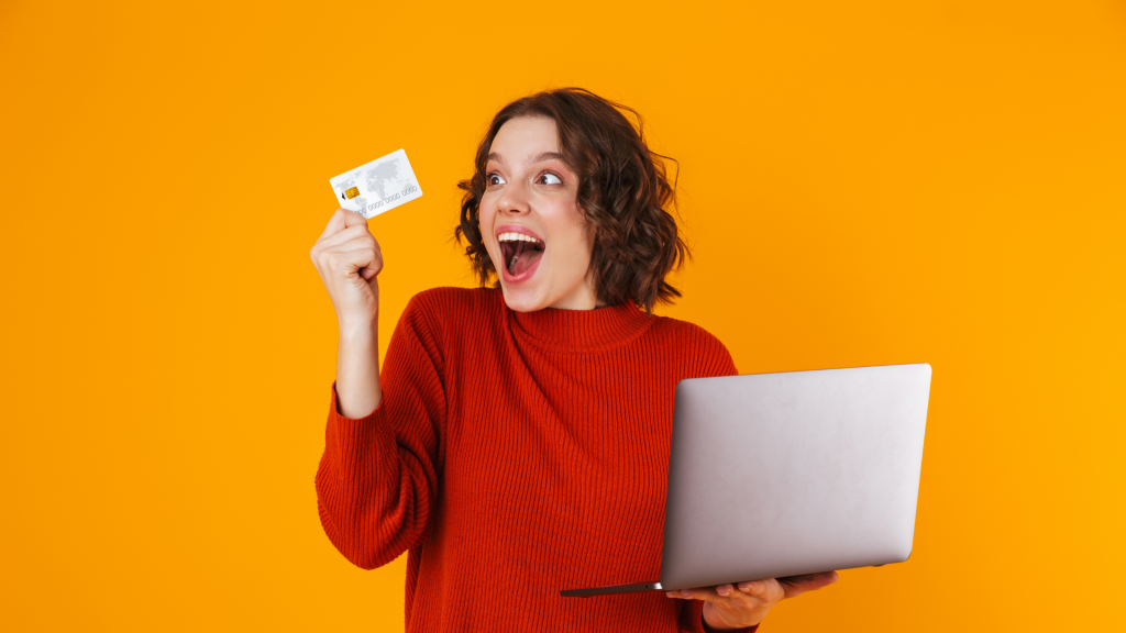 Image of european woman using silver laptop and credit card while standing isolated over yellow background (apply Wells Fargo Active Cash℠ card)