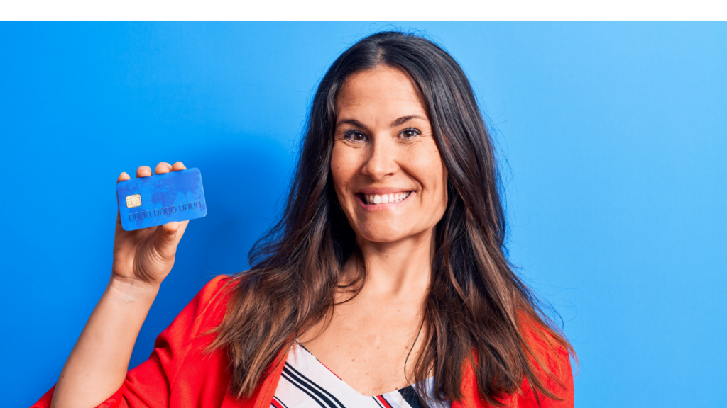 Young beautiful brunette businesswoman holding credit card over isolated blue background looking positive and happy standing and smiling with a confident smile showing teeth