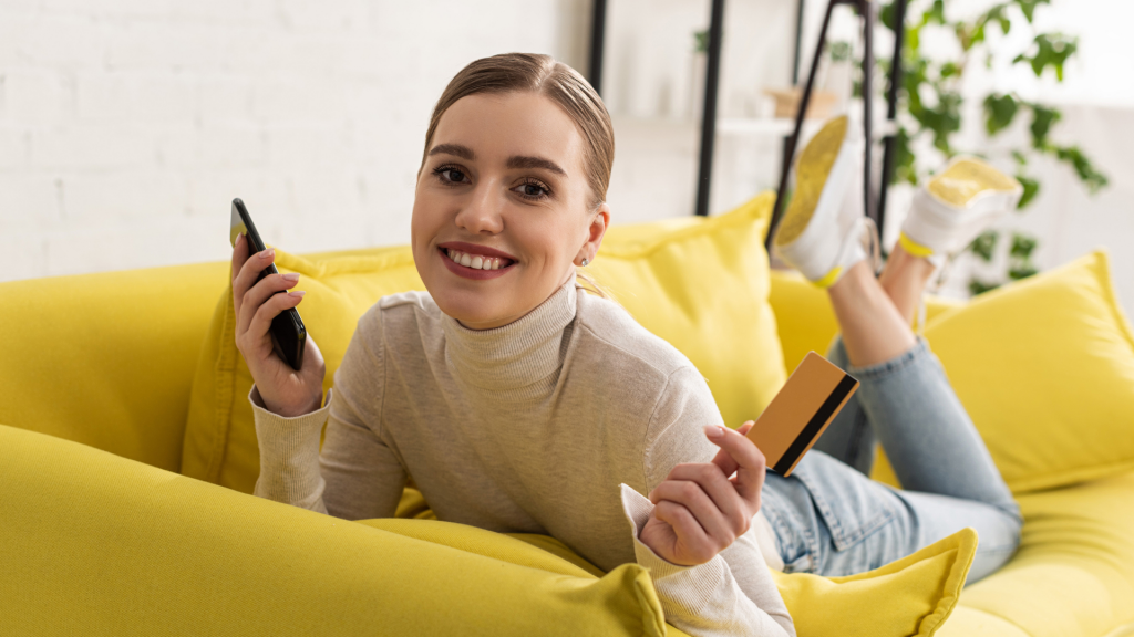 Smiling woman holding smartphone and credit card while lying on sofa (PREMIER Bankcard® Mastercard® credit card)