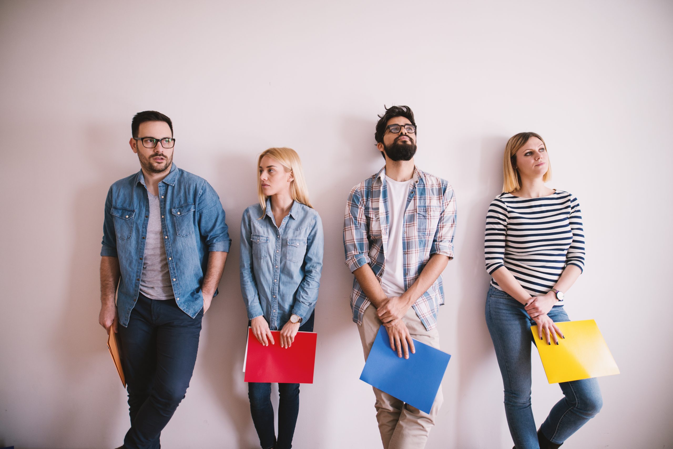 Group of young people leaning against the wall bored before a job interview with folders in hands in the waiting room.