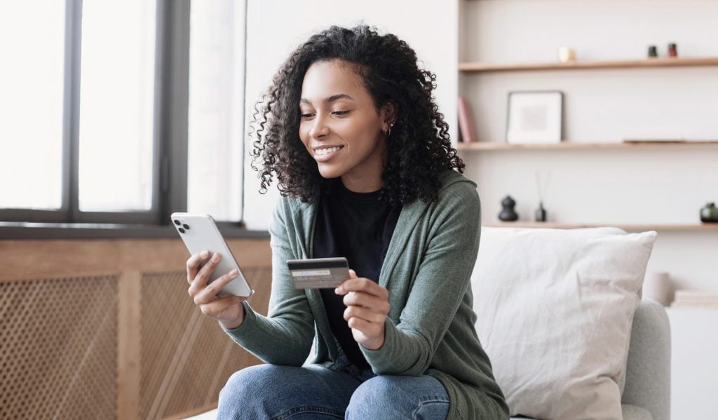 Woman holding credit card and using smartphone at home, business