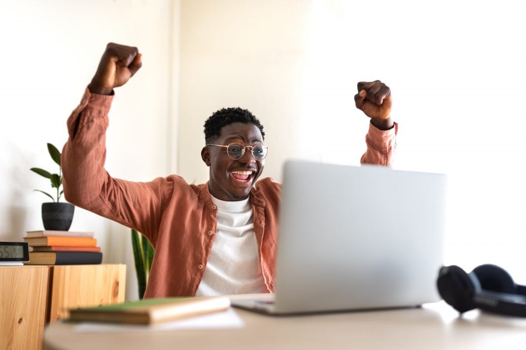 Happy young black male celebrating success with arms raised up in front of laptop. Achievement concept.