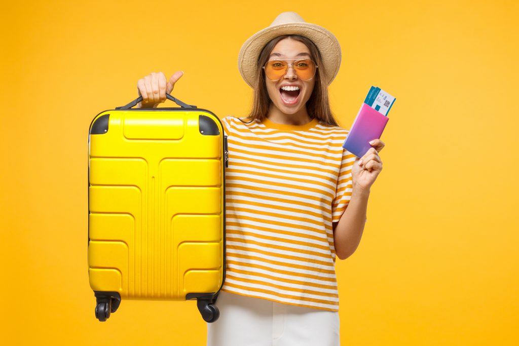 Happy European girl with suitcase and airplane tickets isolated on yellow background excited about trip