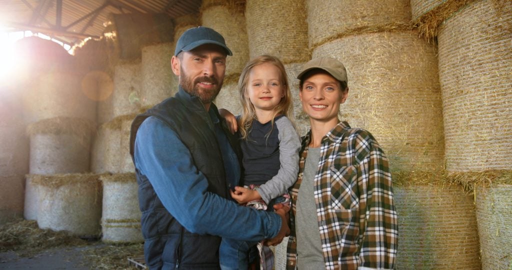 Portrait of happy Caucasian family of farmers with kid standing at barn with hay stocks and smiling to camera. Handsome father, beautiful mother and small pretty daughter at countryside farm. Village.