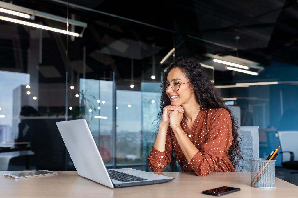 Happy and smiling hispanic businesswoman typing on laptop, office worker with curly hair and glasses happy with achievement results, at work inside office building