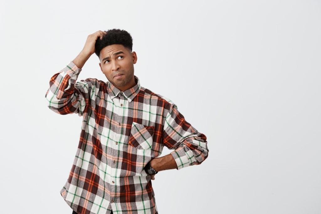 Portrait of young confused dark-skinned handsome man with afro hairstyle in checkered shirt holding head with hand, looking aside with satisfied expression, don't know what to do with debts.