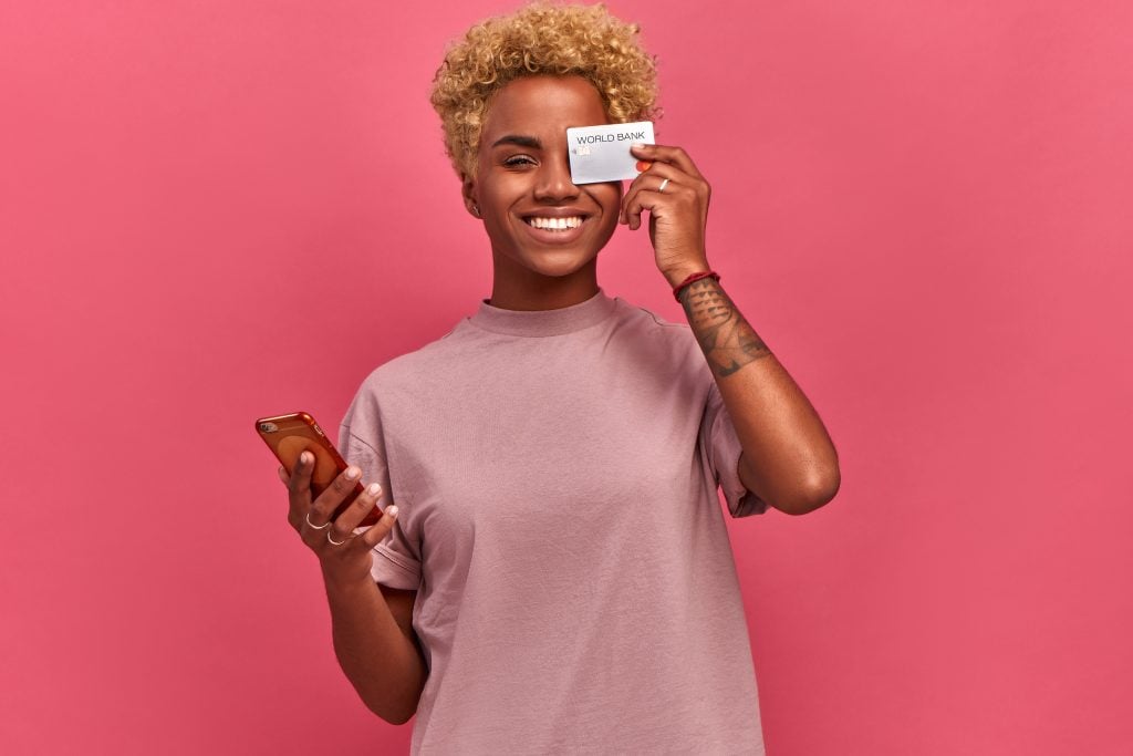 Young dark-skinned woman freelancer holding a bank card and phone on a pink background.