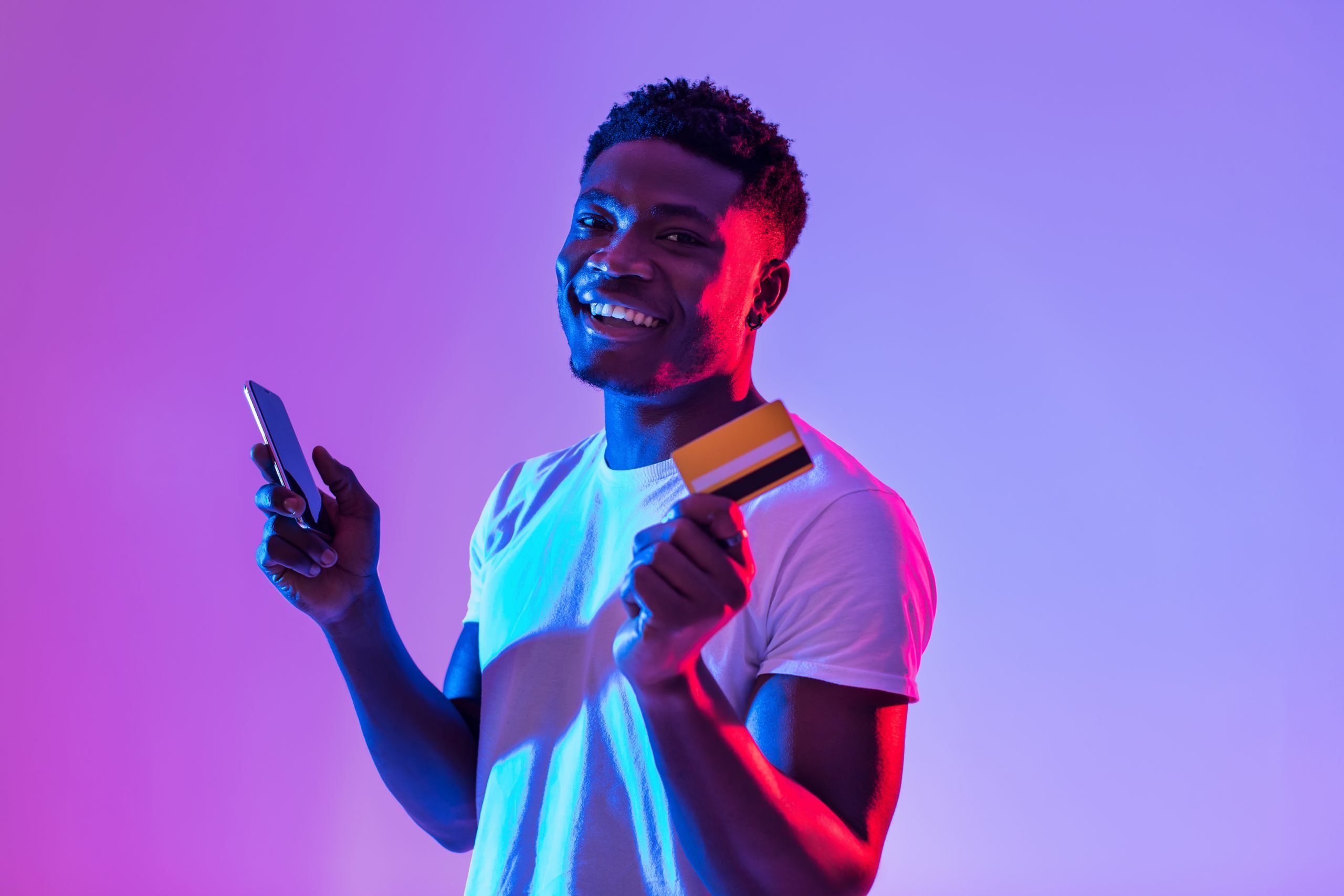 Young black man holding cellphone and credit card, using banking or shopping mobile app in neon light