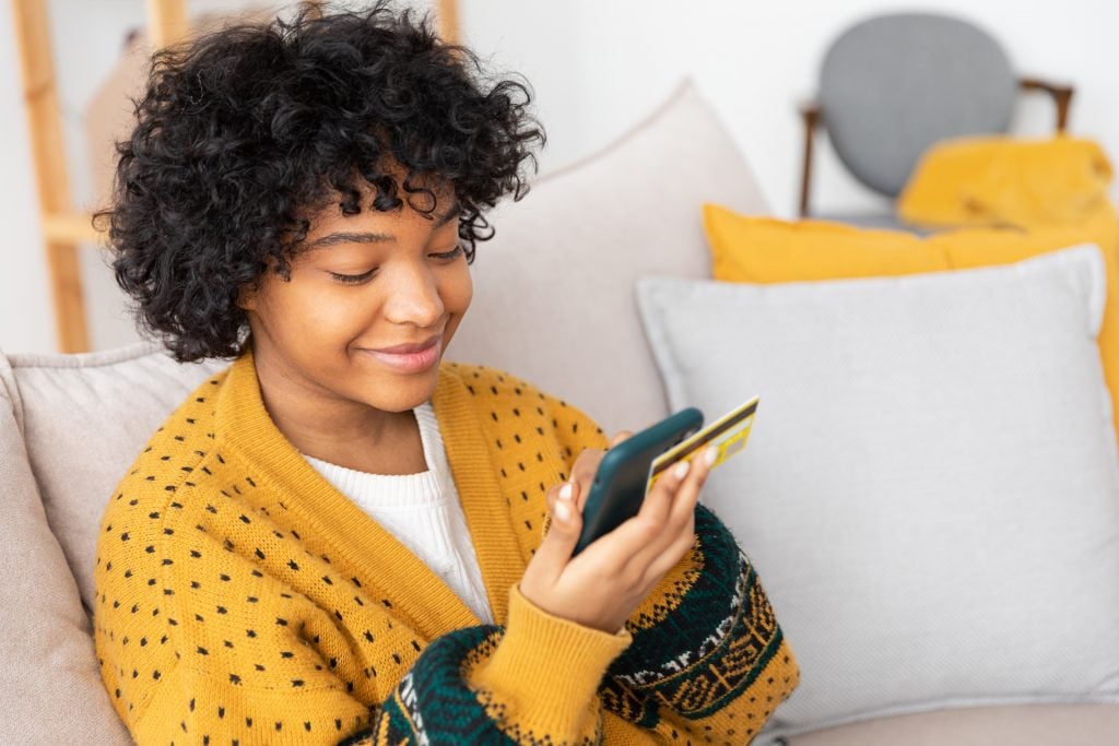 African american woman shopping online holding smartphone paying with gold credit card. Girl sitting at home buying on Internet enter credit card details. Online shopping ecommerce delivery service.