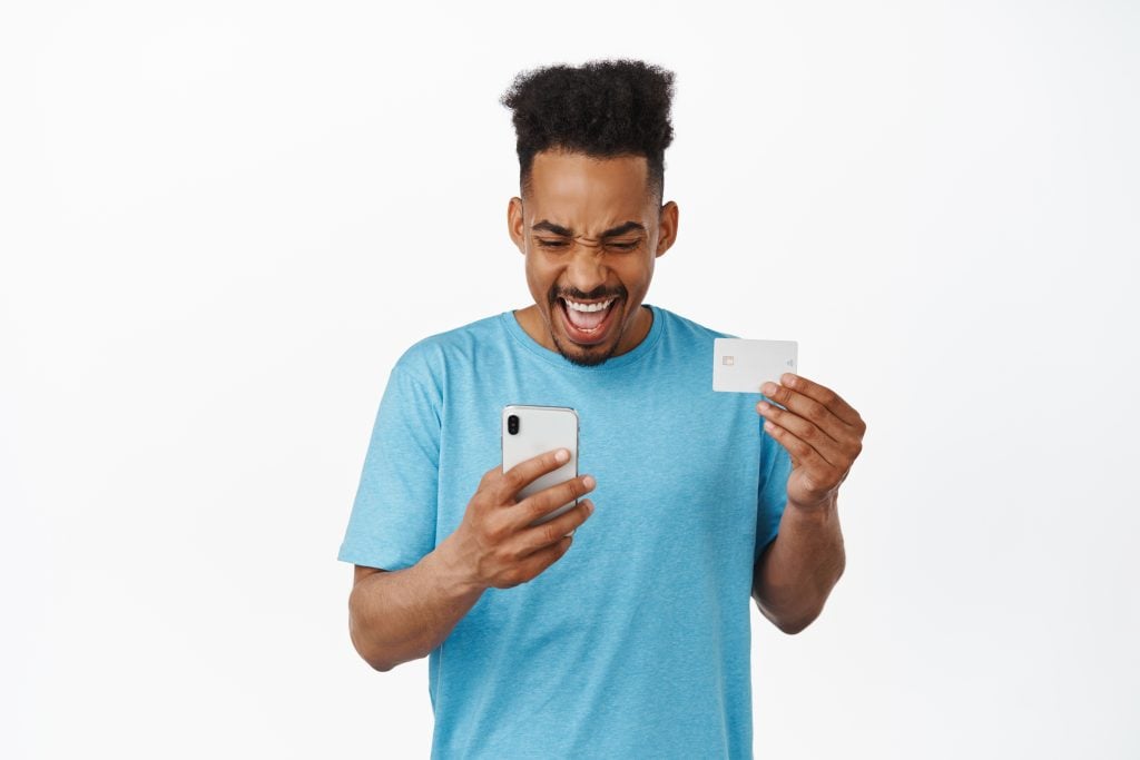 Cheerful african american man looking at smartphone and credit card, paying, shopping online, smiling excited, making purchase, standing against white background