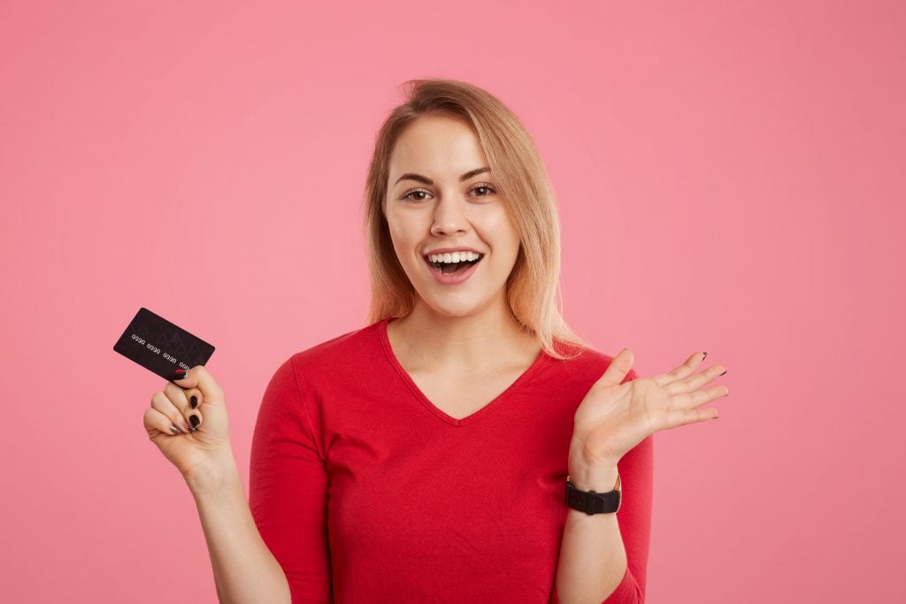 Happy excited blonde female holds plastic card, doesn`t expect to recieve salary, going to make payments, looks joyfully into camera, isolated over pink background. Woman with credit card in hands