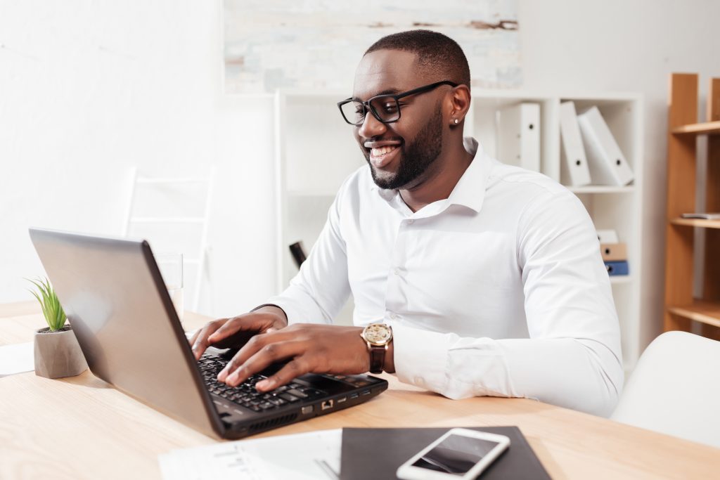 portrait-smiling-african-american-businessman-white-shirt-eyewear-sitting-working-his-laptop-office-isolated