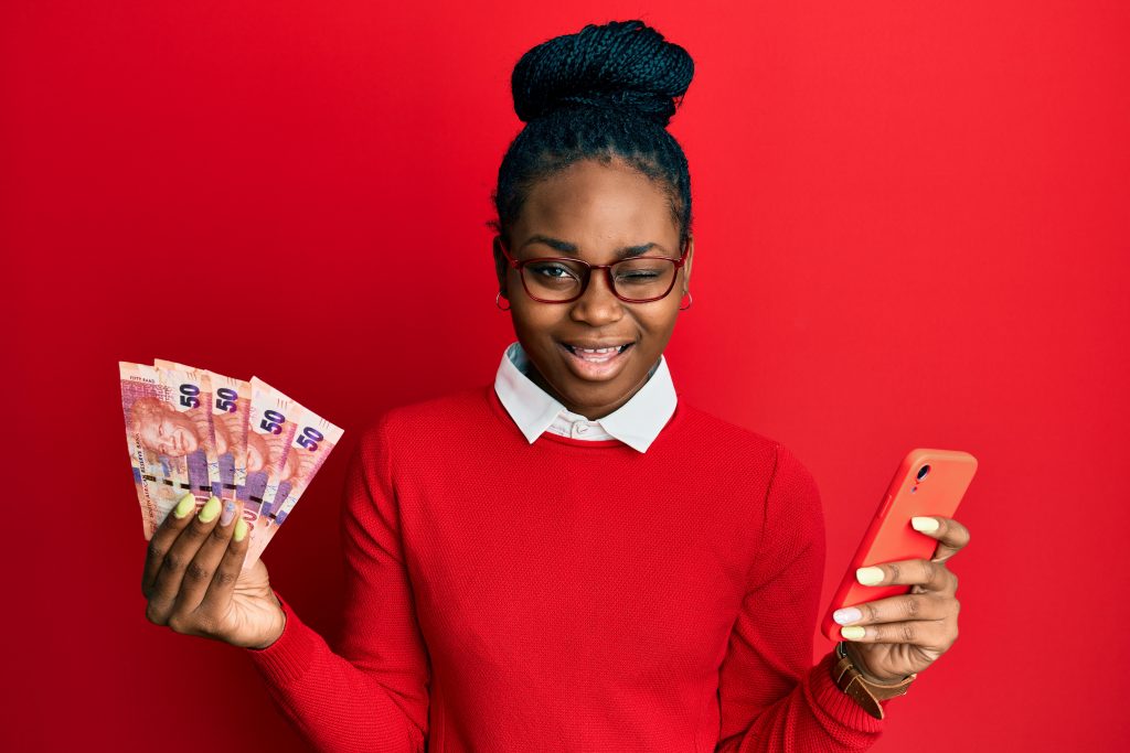 Young african american woman using smartphone holding south africa rand banknotes winking looking at the camera with sexy expression, cheerful and happy face.