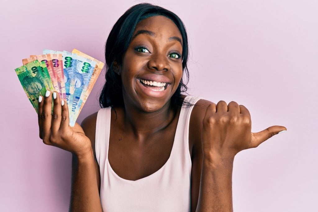 Young african american woman holding south african rand banknotes pointing thumb up to the side smiling happy with open mouth