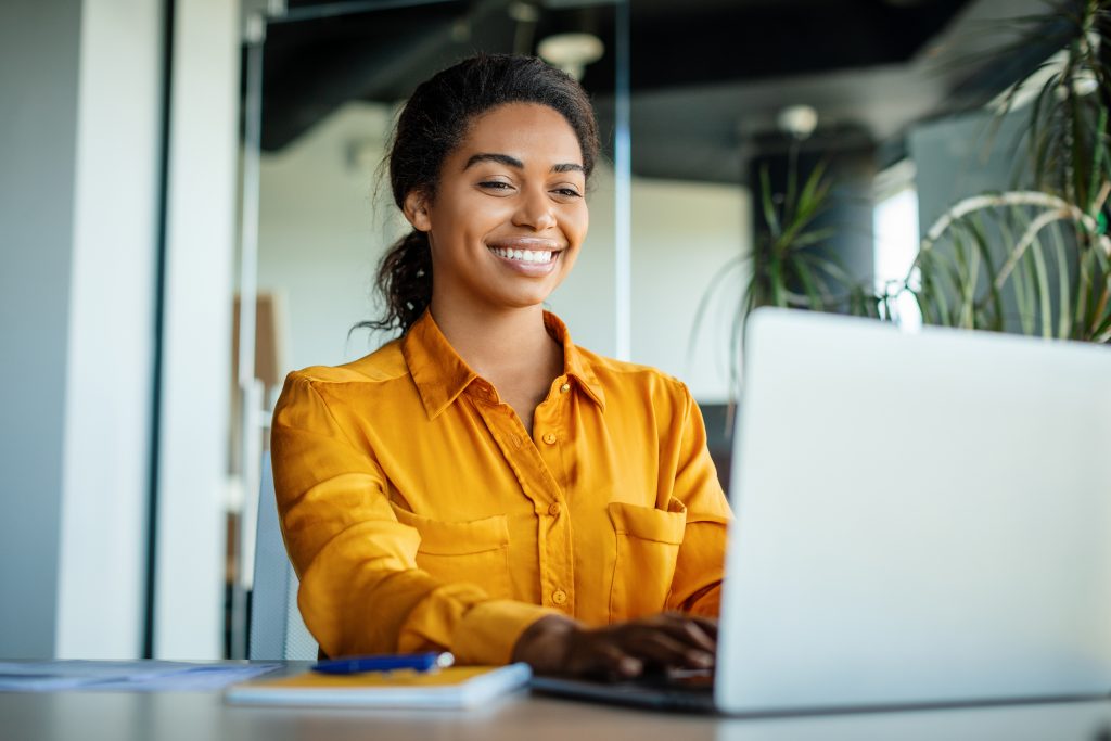 Happy black businesswoman using laptop typing and working online while sitting at workplace in office, copy space