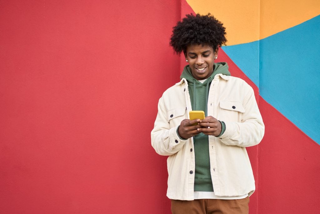 Smiling young African guy standing at colorful wall outdoors using cell phone.