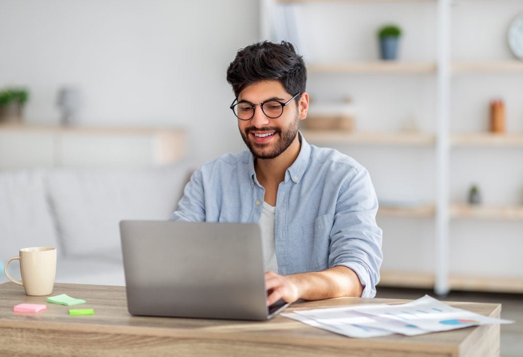 Portrait of happy arab freelancer man sitting at desk with laptop computer at home office, looking and smiling at screen