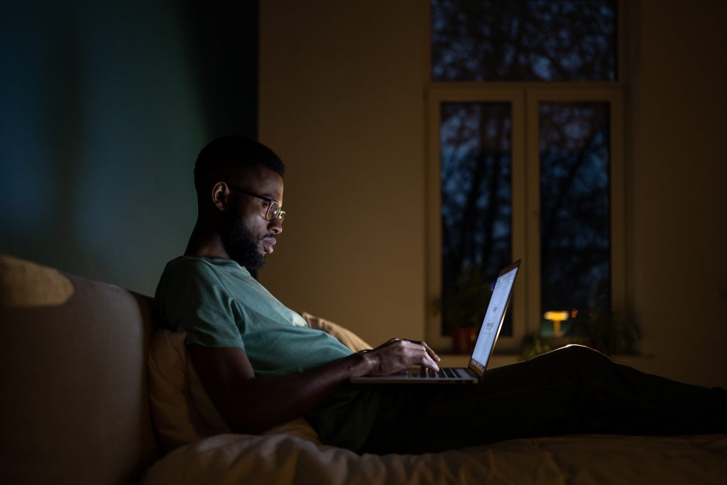 Concentrated African American programmer man lying on bed work on laptop at night.