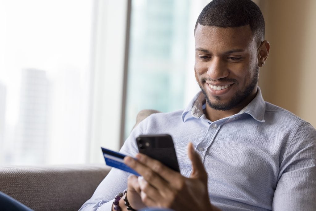 Happy millennial man using credit card, ecommerce app on mobile phone for payment, online order, shopping, buying, sitting on home couch, typing, smiling, laughing