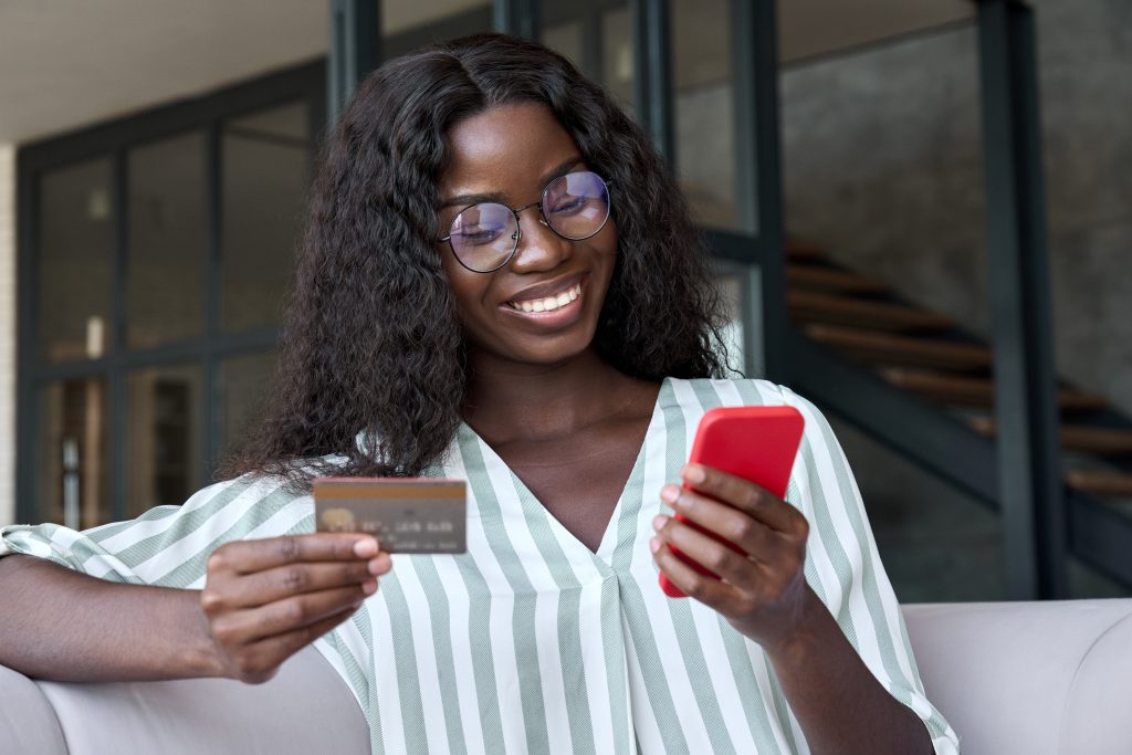 Happy young black woman shopper holding credit card using phone buying online.